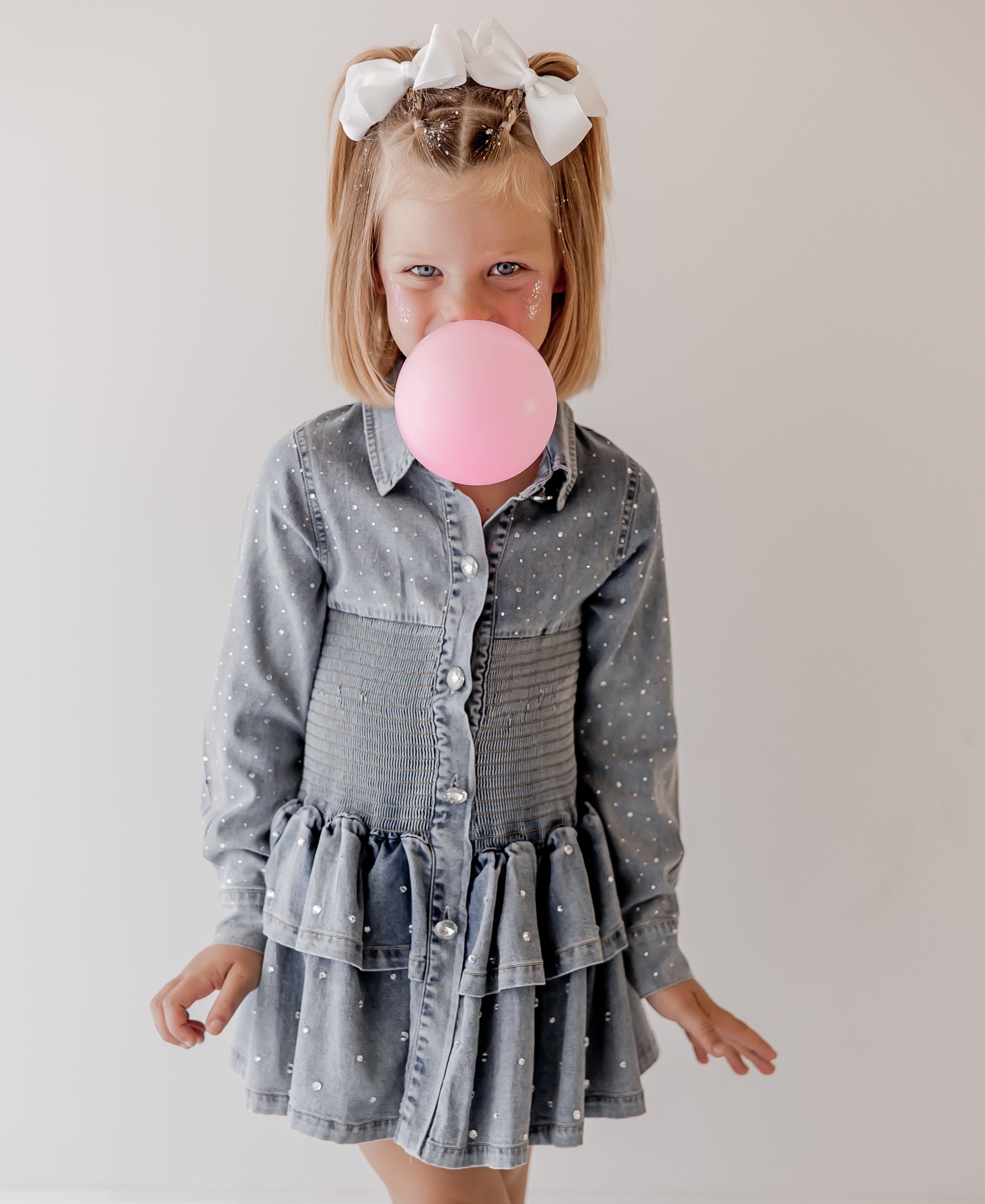 Young girl in a denim dress blowing a pink bubble with a white background