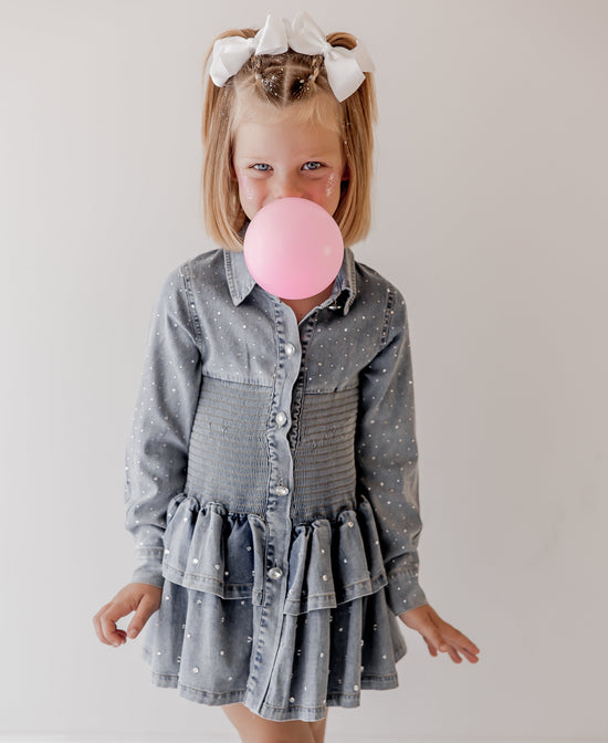 Young girl in a denim dress blowing a pink bubble with a white background