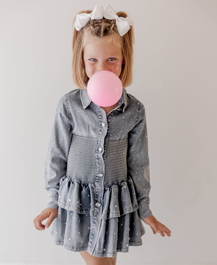Young girl in a denim dress blowing a pink bubble with a white background